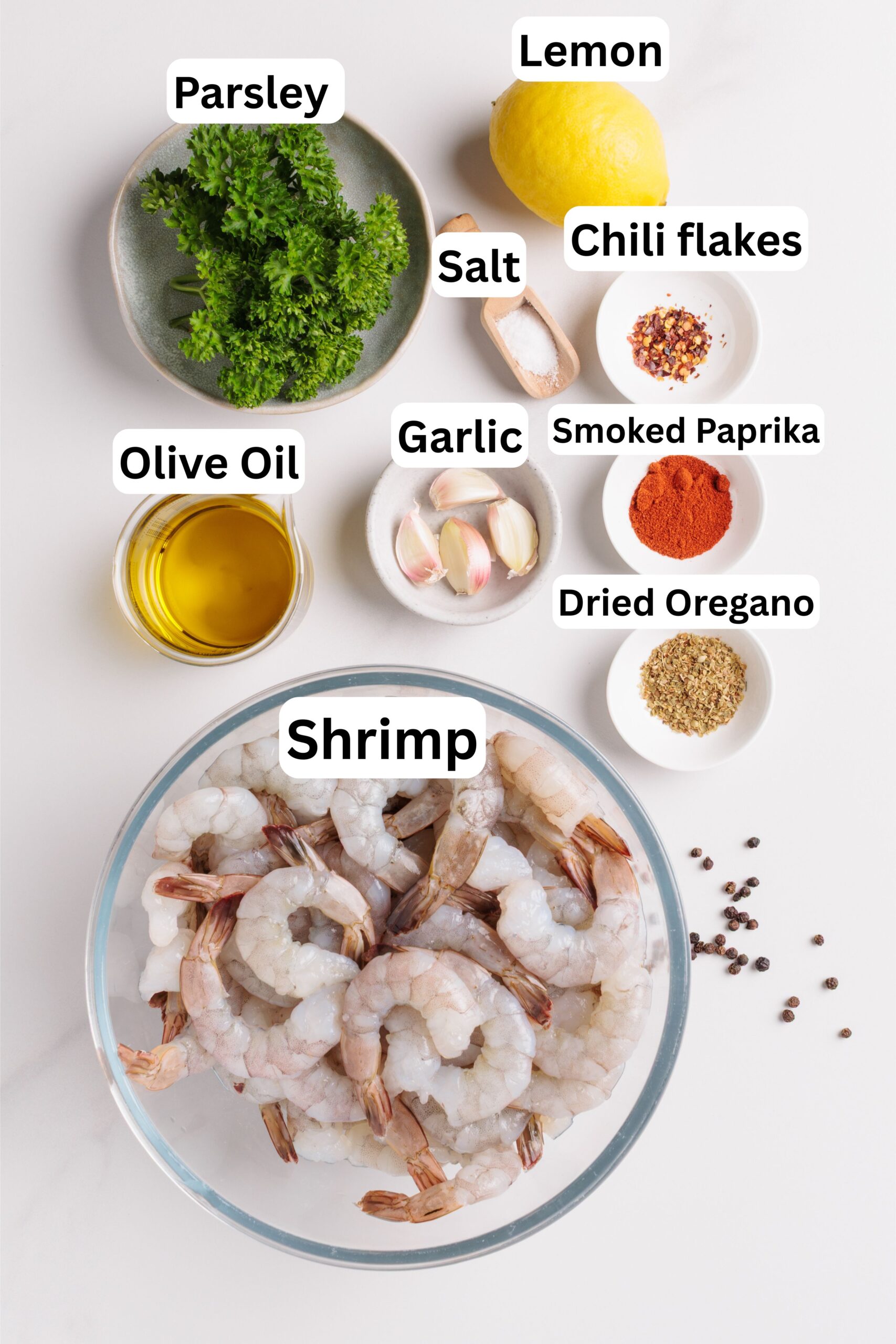 A bowl of raw shrimp surrounded by small bowls of olive oil, parsley, salt, garlic, chili flakes, smoked paprika, dried oregano, and a lemon, all labeled with text.
