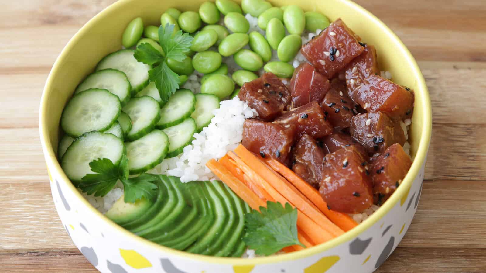 A ahi tuna poke bowl with sliced cucumber, avocado, edamame, shredded carrots, white rice, marinated tuna, and parsley garnish in a yellow and white bowl on a wooden surface.