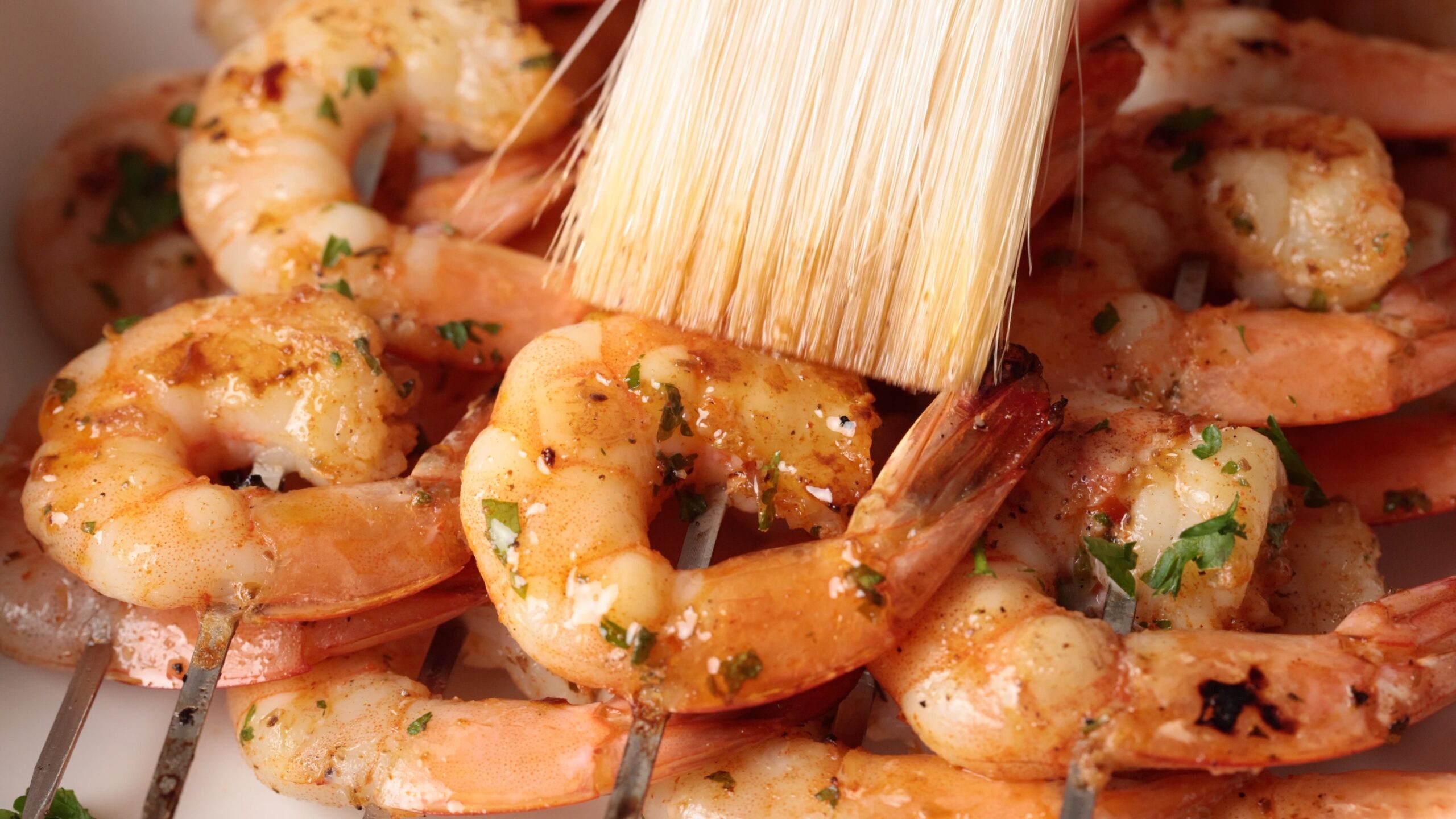 A close-up of grilled shrimp on skewers being brushed with a marinade or sauce. The shrimp are garnished with chopped herbs and appear juicy and flavorful.