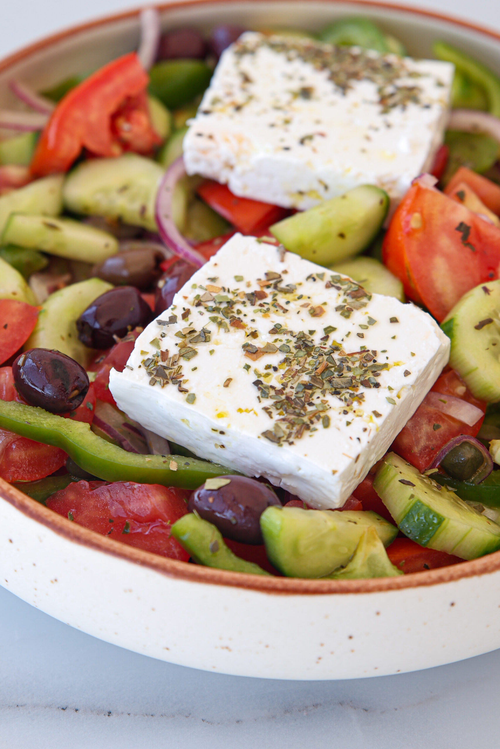 A bowl of Greek salad with tomato, cucumber, olives, red onion, green pepper, and two slices of feta cheese topped with herbs.