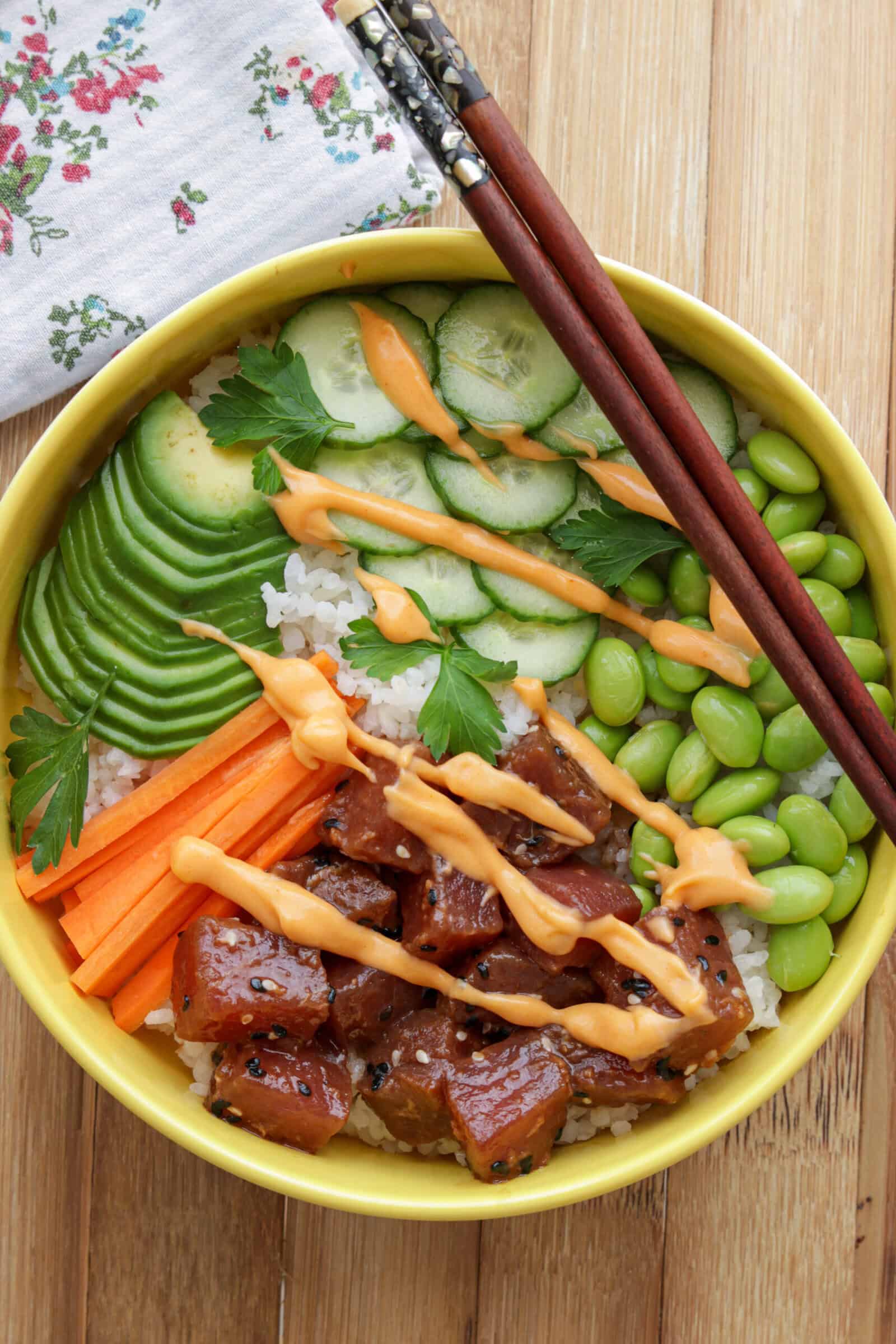 A bowl of tuna poke bowl with rice, cubed marinated ahi tuna fish, sliced avocado, cucumber, edamame, carrot sticks, parsley, and orange sauce, with chopsticks on top, on a wooden surface.