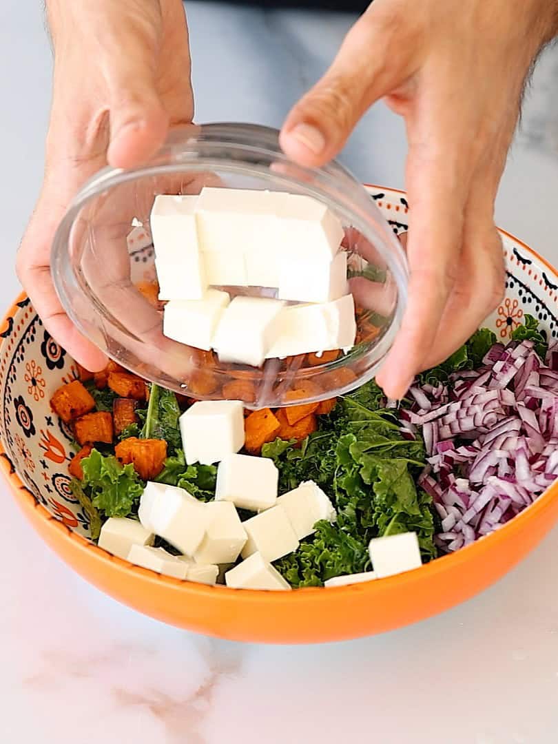 A person adds cubes of white cheese from a small bowl into a larger orange bowl containing chopped kale, red onion, and roasted sweet potatoes.