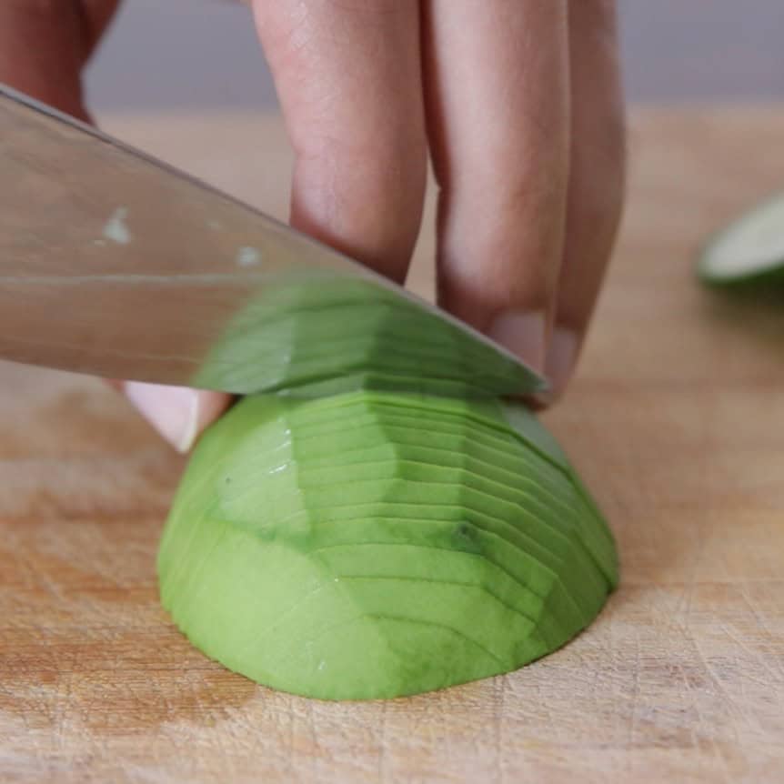 A hand slices an avocado half on a wooden cutting board using a large kitchen knife.