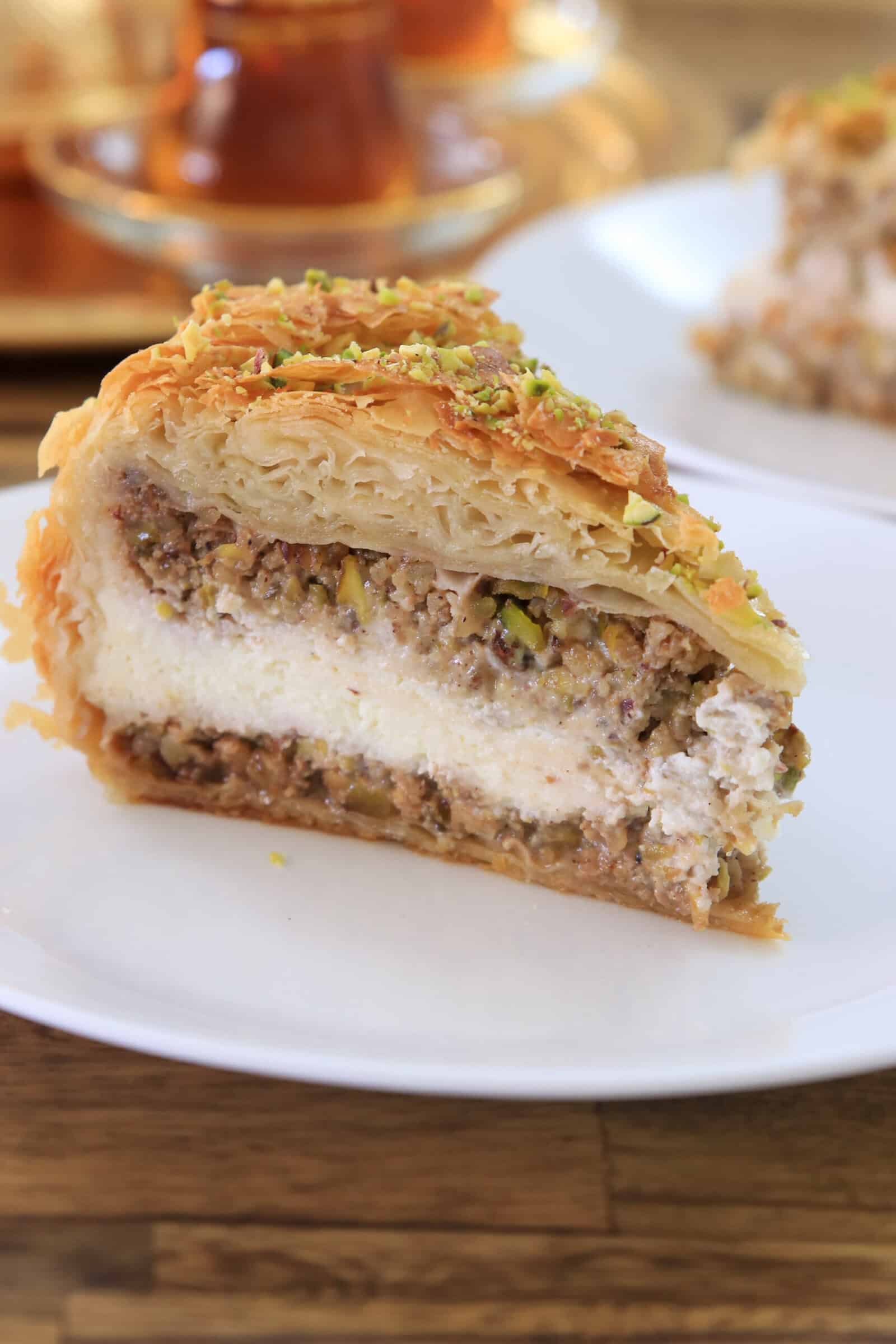 A close-up of a slice of baklava cheesecake with a nut and cream filling, topped with finely chopped pistachios, on a white plate. A blurred tea glass and another plate are in the background.