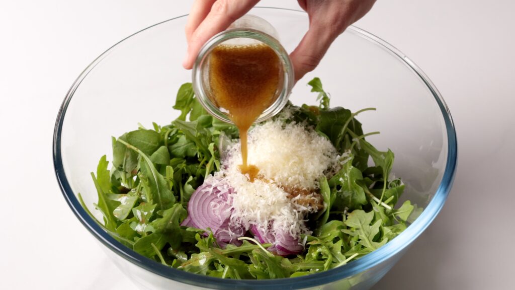 A hand pours brown vinaigrette dressing from a small jar onto a bowl of arugula, red onion slices, and grated cheese in a clear glass bowl.