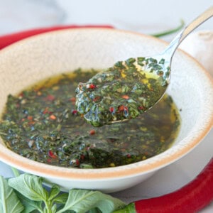 A spoonful of chimichurri sauce with basil chopped herbs and oil is held over a bowl, surrounded by fresh basil leaves and a red chili pepper.