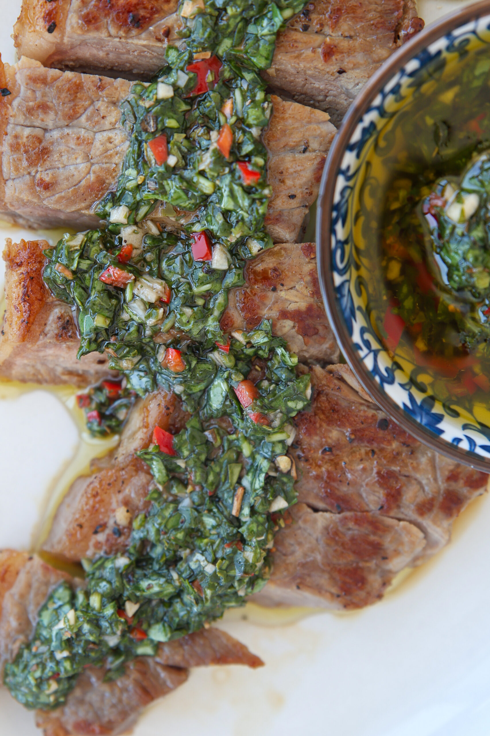 Sliced grilled steak topped with vibrant green chimichurri sauce, served with a small bowl of extra chimichurri on the side.