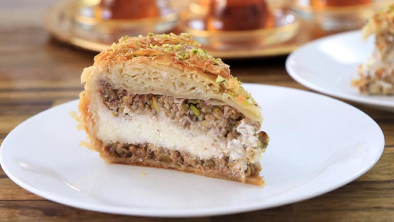 A slice of layered baklava cheesecake with nuts and creamy filling sits on a white plate, with more dessert and tea glasses blurred in the background.