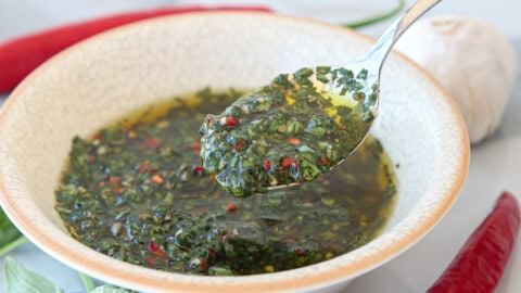 A spoon scoops up vibrant green basil chimichurri sauce from a bowl, with visible herbs and red pepper flakes. Garlic and red chili peppers are blurred in the background.