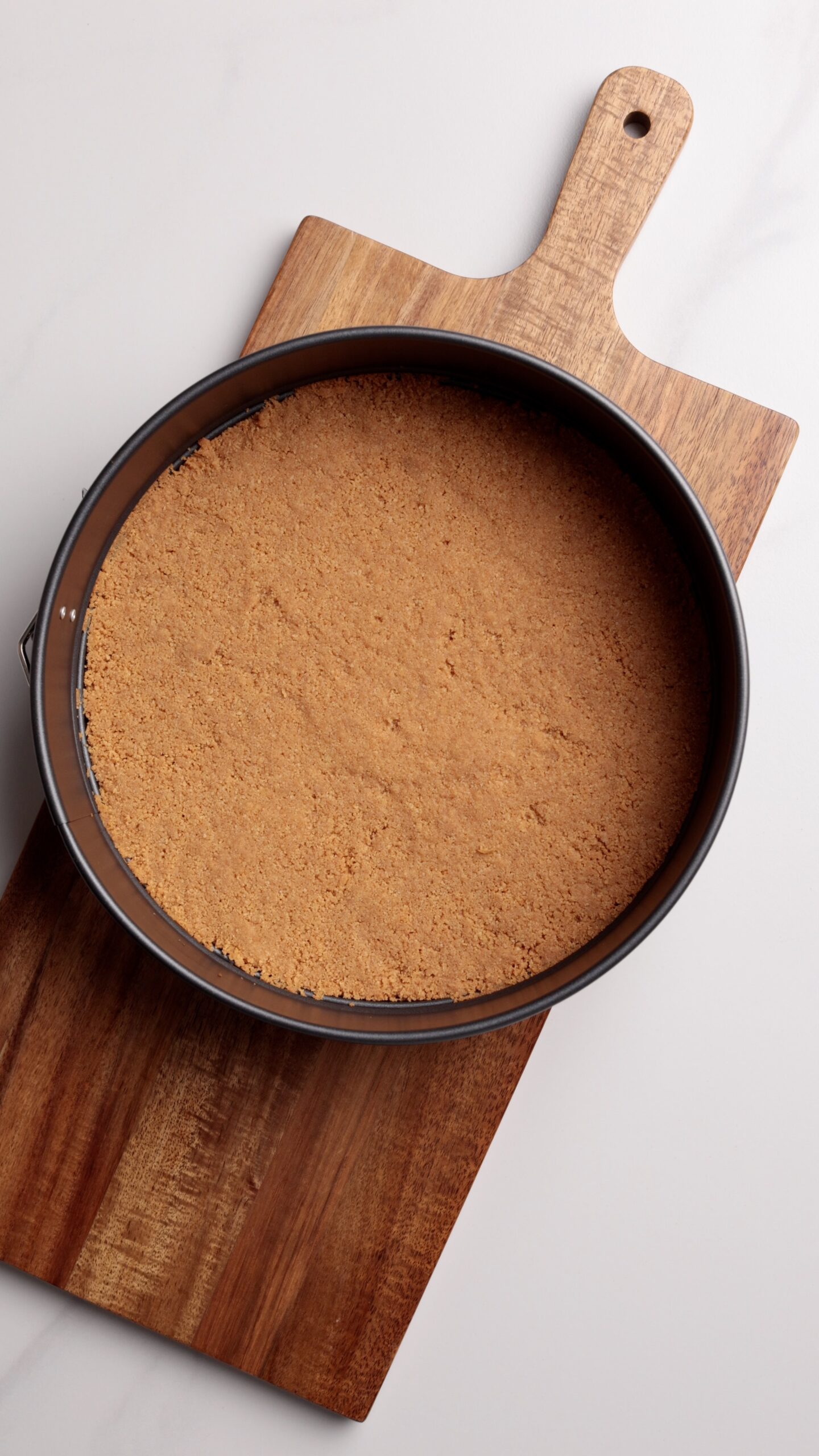 A round, springform pan filled with a pressed, golden-brown graham cracker crust sits on a wooden cutting board against a light background.