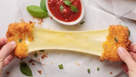 Crispy Fried Mozzarella (Sticks, Squares, Balls & More) Two hands pull apart a fried mozzarella stick, stretching melted cheese between them. A bowl of marinara sauce and basil leaves sit nearby on a white surface.