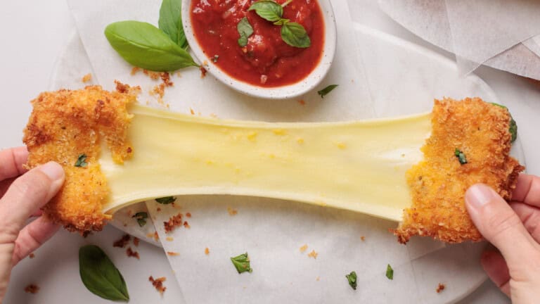Two hands pull apart a fried mozzarella stick, stretching melted cheese between them. A bowl of marinara sauce and basil leaves sit nearby on a white surface.