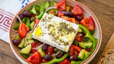 A bowl of Greek salad with cucumbers, tomatoes, green peppers, red onions, olives, and a slab of feta cheese topped with herbs and olive oil. Bread slices are visible on the side.