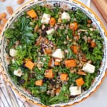 A colorful salad with kale, lentils, roasted sweet potatoes, feta cheese cubes, almonds, red onions, and raisins in a patterned bowl on a table.