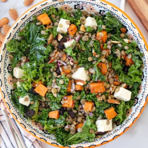 A colorful salad with kale, lentils, roasted sweet potatoes, feta cheese cubes, almonds, red onions, and raisins in a patterned bowl on a table.