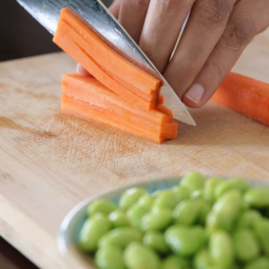 A hand slices carrot sticks with a knife on a wooden cutting board. A small plate with shelled edamame beans is visible in the foreground.