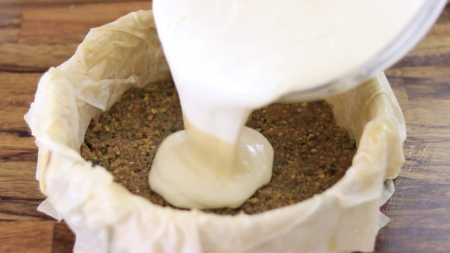 A creamy batter is poured into a baking pan lined with parchment paper over a pistachio crust, placed on a wooden surface.