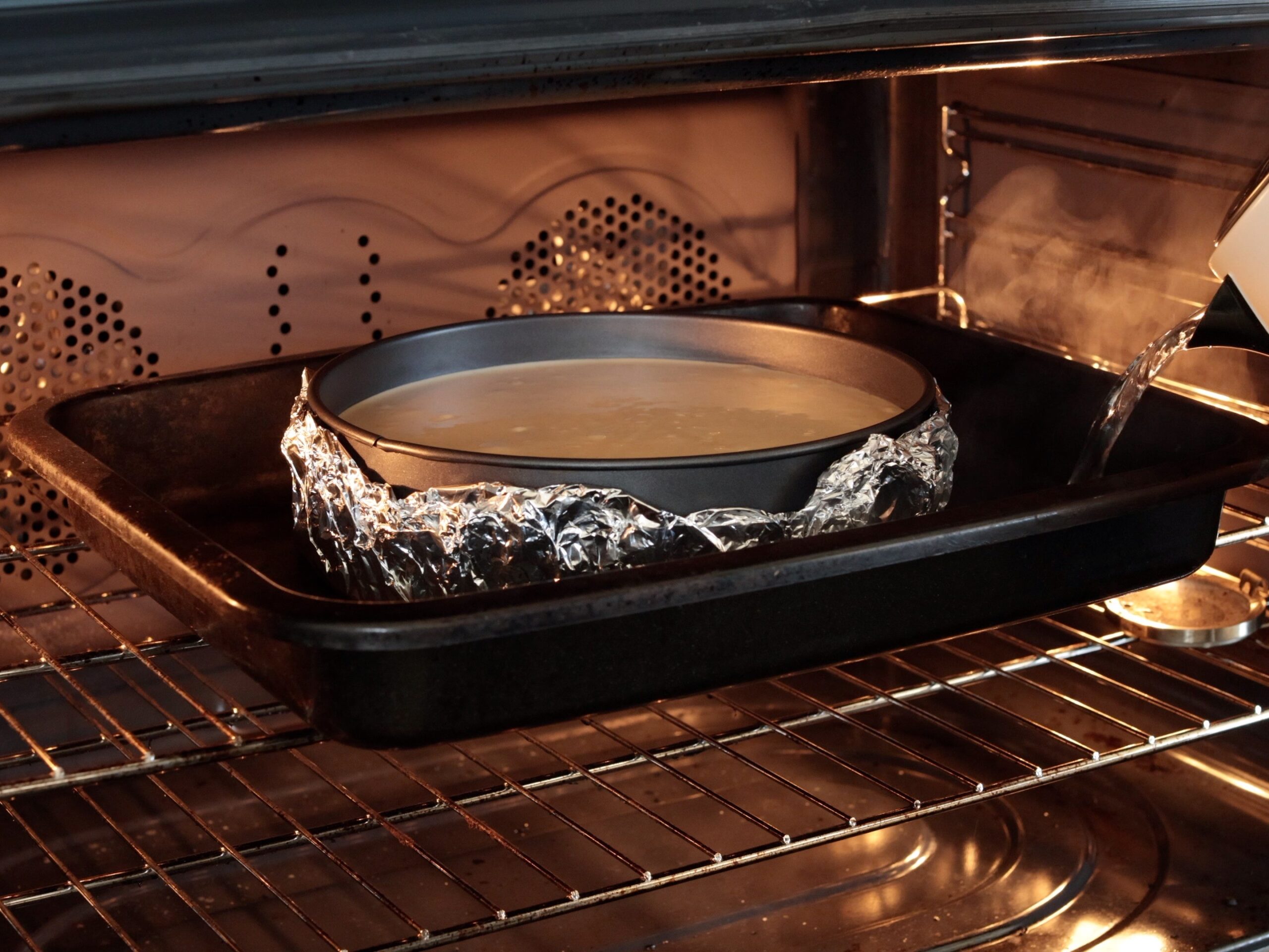 A round cake pan wrapped in foil sits in a larger pan in the oven. Hot water is being poured into the larger pan, creating a water bath for baking.