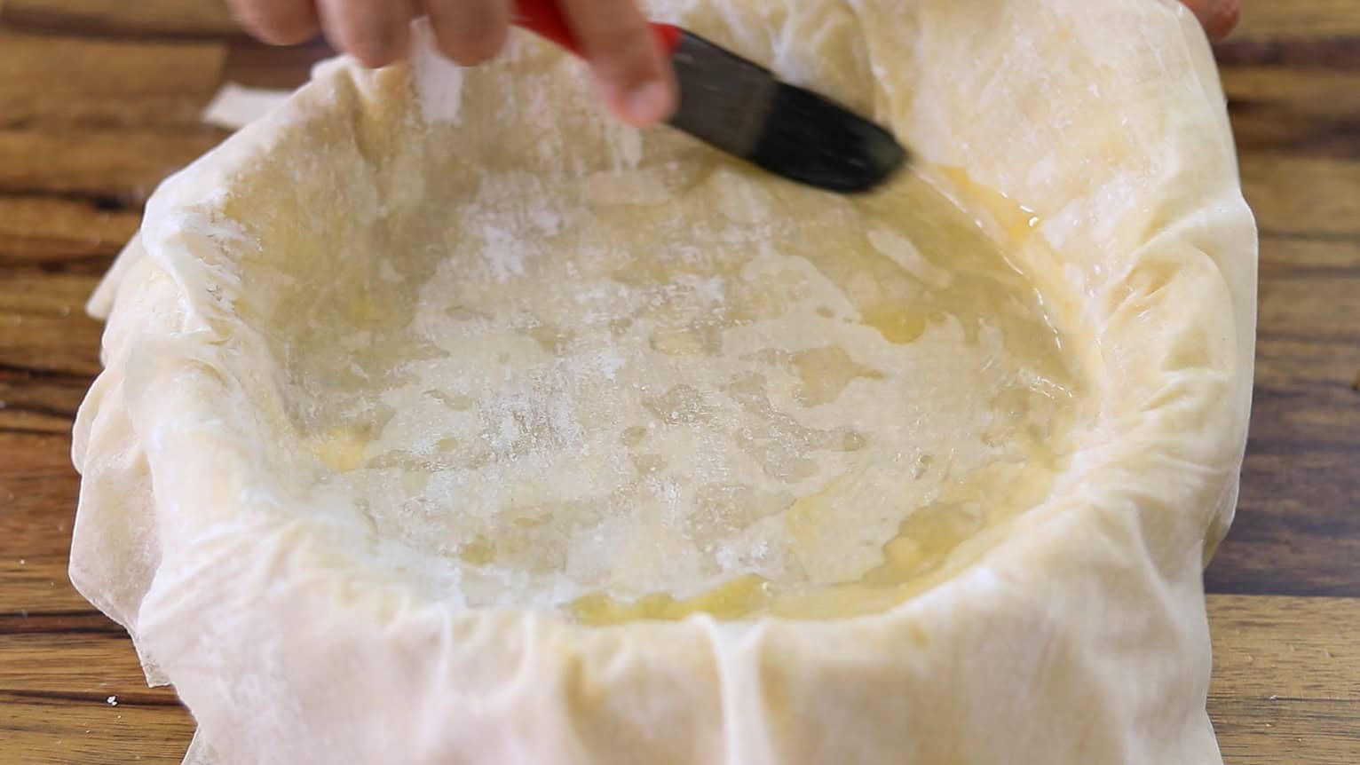 A hand uses a brush to spread butter on a baklava cheesecake lining a round baking dish, with a wooden surface underneath.