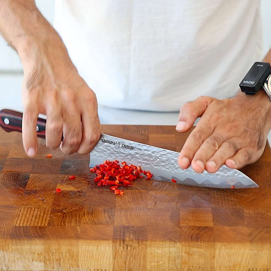 A person finely chops red chili peppers on a wooden cutting board with a large chef’s knife, wearing a black wristwatch.