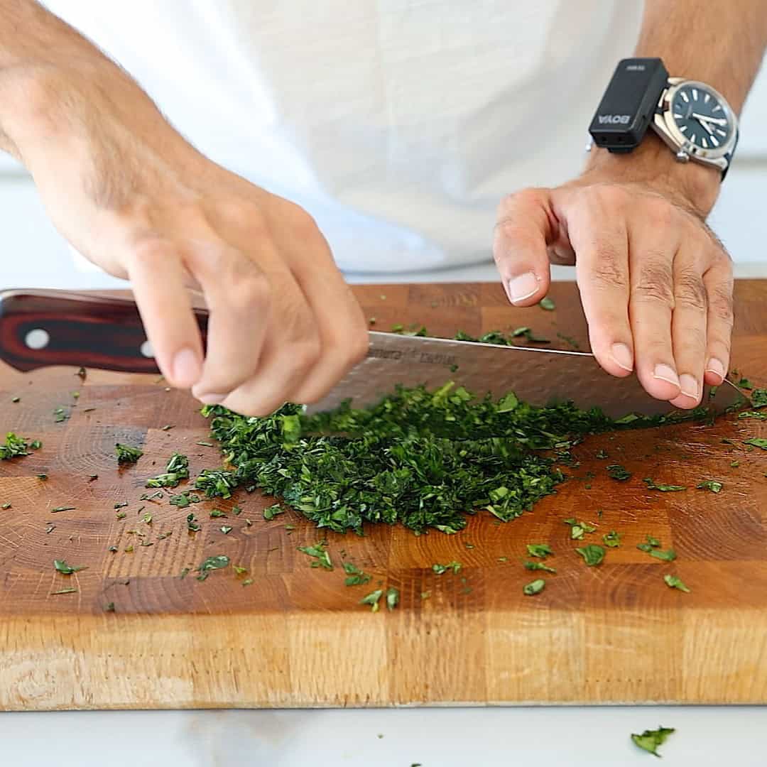 A person chops fresh basil on a wooden cutting board, holding a large knife. They wear a white shirt and a black watch on their left wrist.