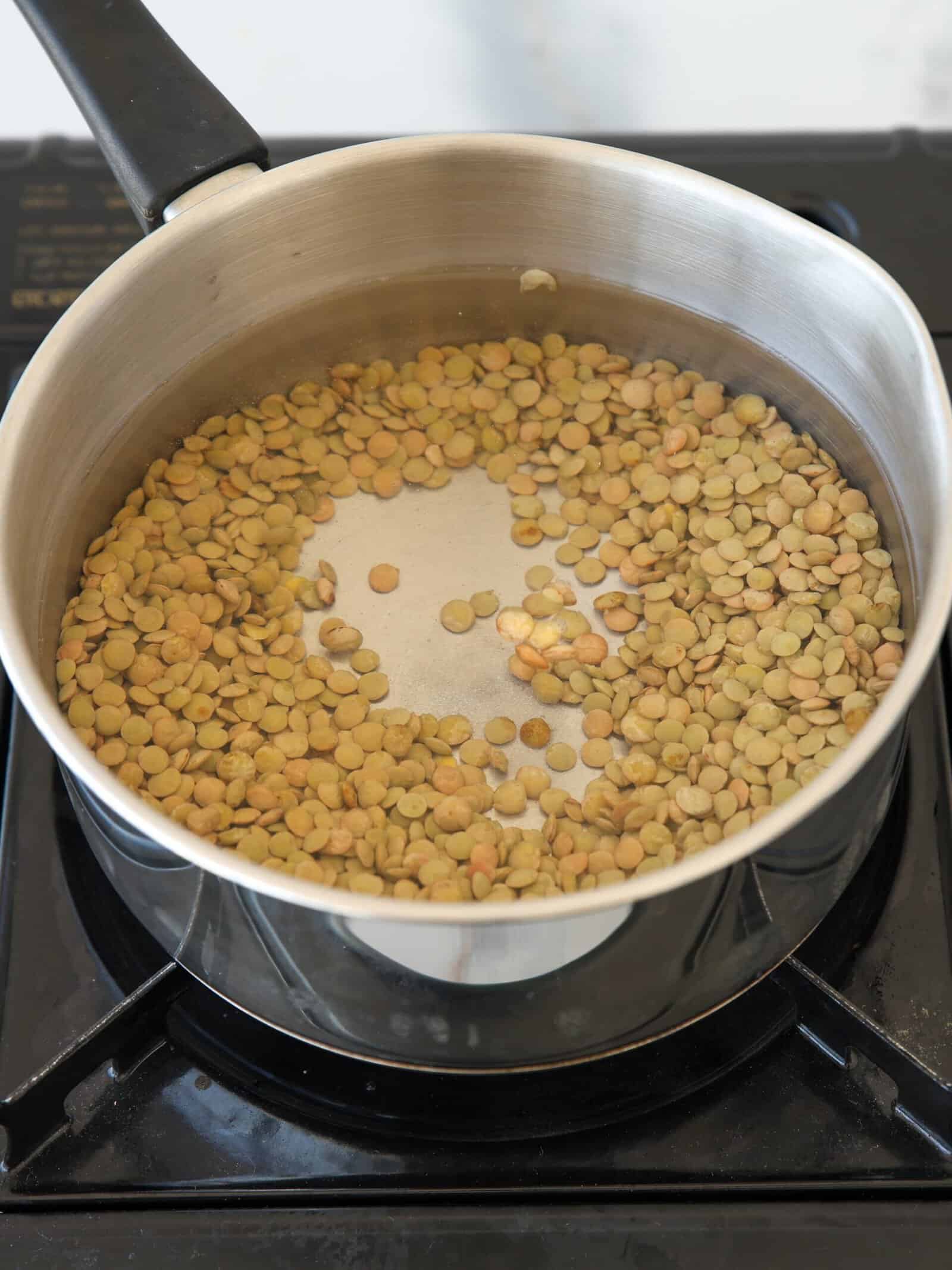 A stainless steel pot with green lentils soaking in water sits on a black stovetop, ready for cooking.