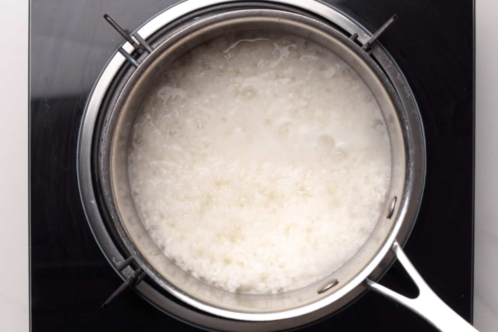 A stainless steel pot of boiling rice with white, starchy liquid sits on a black stove top, viewed from above.