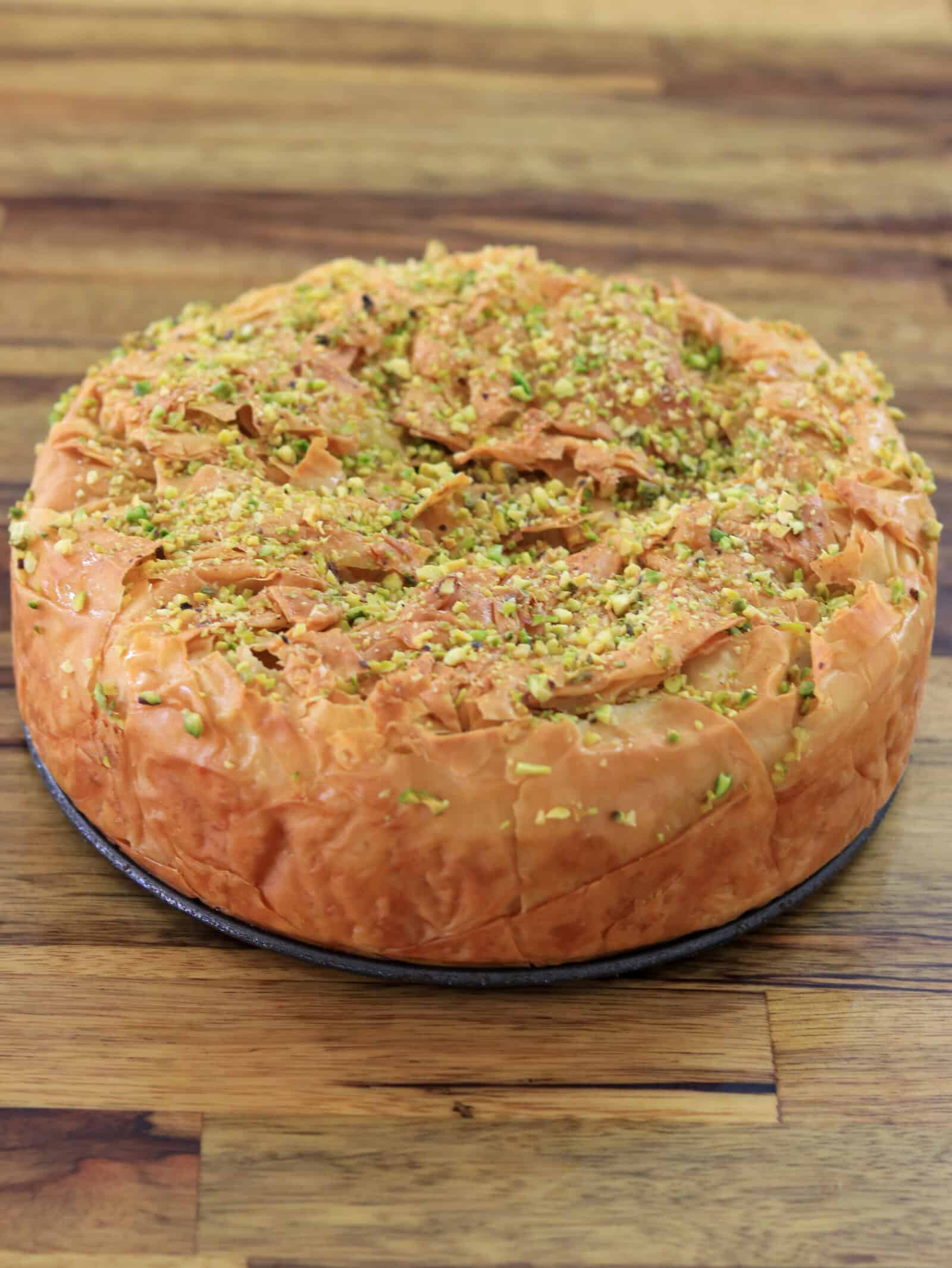 A round, golden-brown baklava cheesecake topped with crushed pistachios sits on a wooden surface. The flaky, layered texture suggests it is a type of Middle Eastern or Mediterranean dessert.