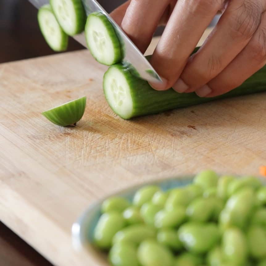 A person slices a cucumber on a wooden cutting board, with a bowl of green edamame beans visible in the foreground.