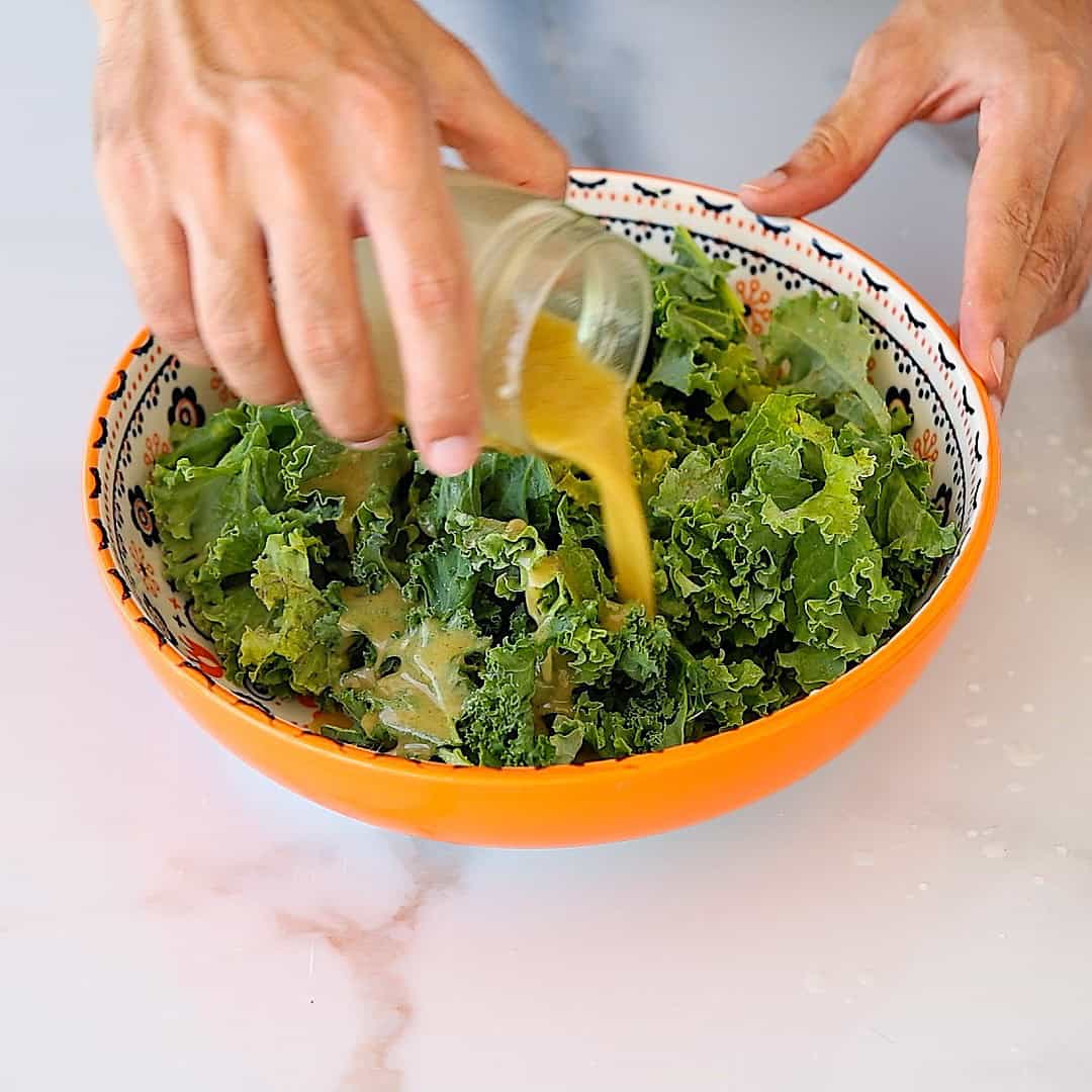 A person pours creamy dressing from a jar over fresh kale in a patterned orange bowl on a white surface.