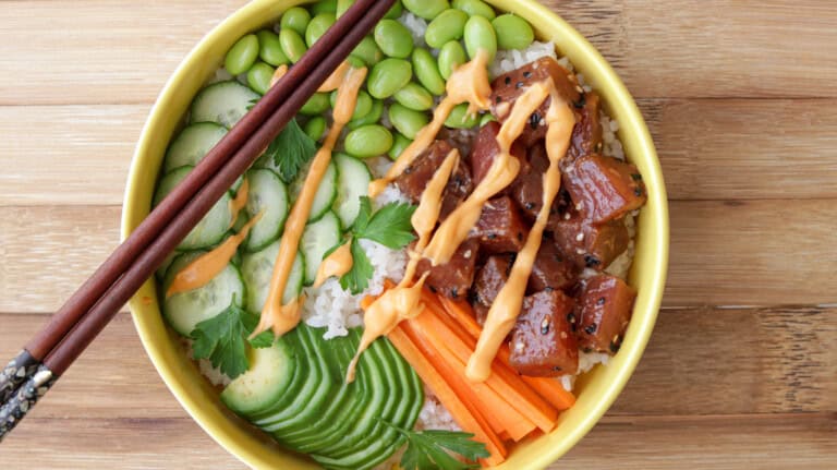 A poke bowl with rice, diced marinated tuna fish, cucumber, avocado, edamame, carrot, parsley, and orange sauce, served with chopsticks on a wooden table.