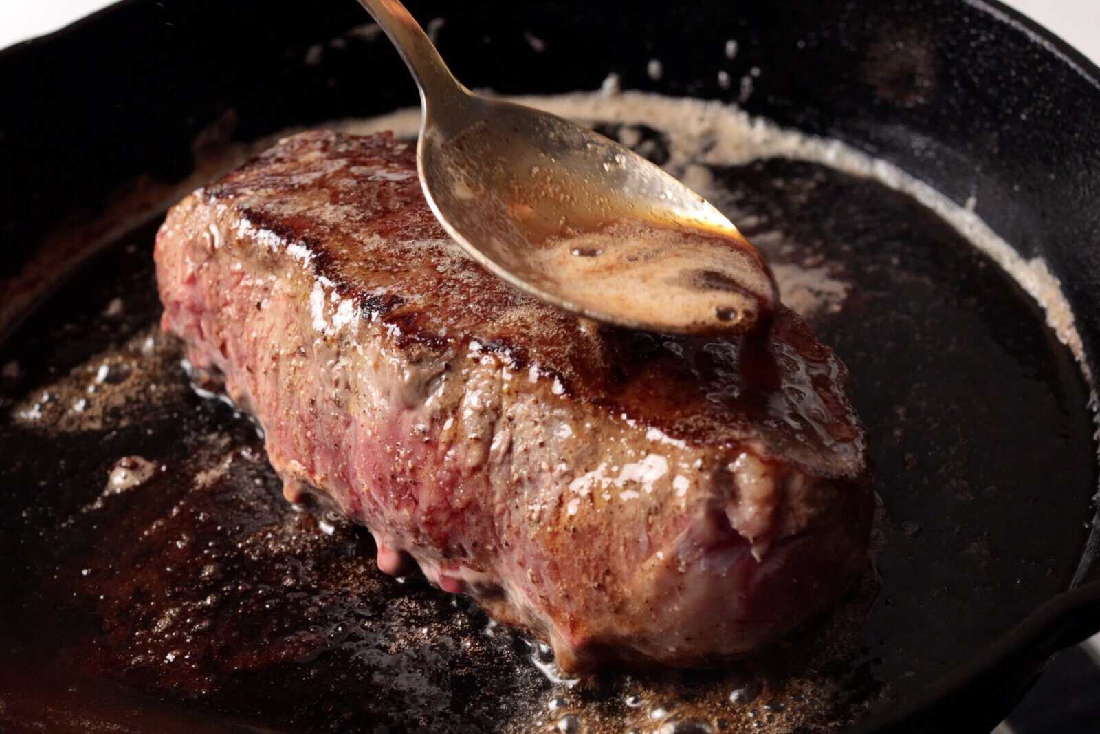 A thick steak is being basted with melted butter using a spoon in a black cast-iron skillet. The steak is searing, with a brown crust forming and juices sizzling around it.