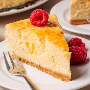 A slice of cheesecake with a golden top sits on a white plate, garnished with fresh raspberries. A silver fork rests beside the dessert. Another slice of cheesecake is visible in the background.