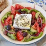 A Greek salad with sliced tomatoes, cucumbers, green peppers, red onions, Kalamata olives, and a large feta cheese block topped with herbs, served in a white bowl.