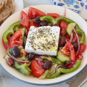 A Greek salad with sliced tomatoes, cucumbers, green peppers, red onions, Kalamata olives, and a large feta cheese block topped with herbs, served in a white bowl.