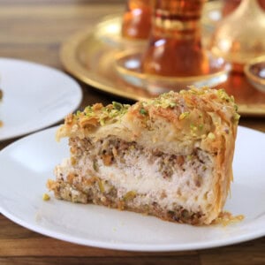 A slice of layered baklava cake filled with nuts and cream, topped with chopped pistachios, sits on a white plate. Decorative tea glasses and a golden tray are blurred in the background.