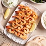 Grilled shrimp skewers garnished with herbs on a white plate with lemon wedges, surrounded by pita bread, rice, and a bowl of creamy dip.