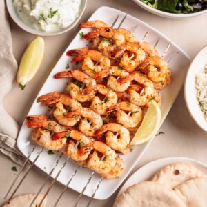 Grilled shrimp skewers garnished with herbs on a white plate with lemon wedges, surrounded by pita bread, rice, and a bowl of creamy dip.