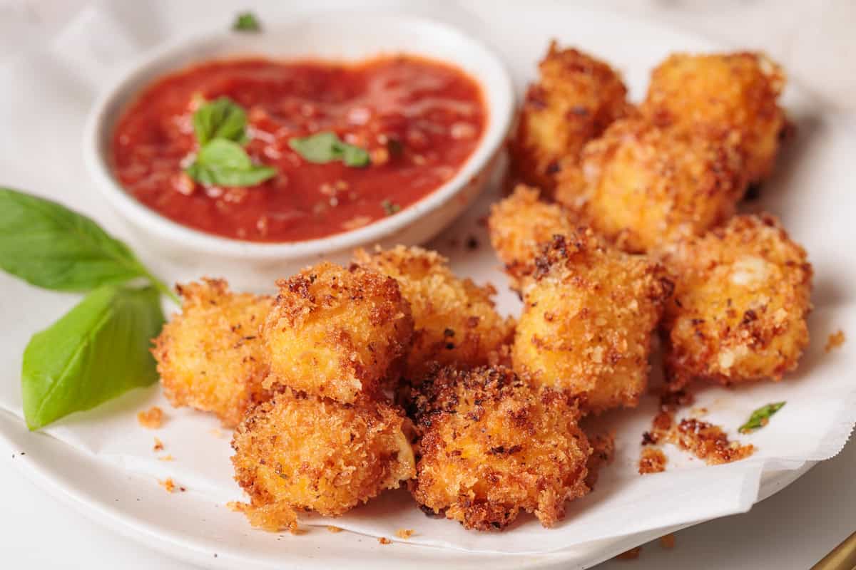 Crispy, golden-brown fried mozzarella balls on parchment paper, served with a bowl of red marinara sauce and garnished with fresh basil leaves.