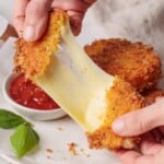 Hands pull apart a crispy, golden breaded mozzarella squares, revealing gooey, stretchy melted cheese inside. A small bowl of marinara sauce and a basil leaf sit nearby on a white surface.
