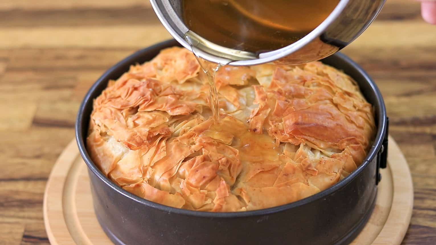 A golden, flaky baklava cheesecake in a round baking pan is being drizzled with syrup from a saucepan on a wooden surface.