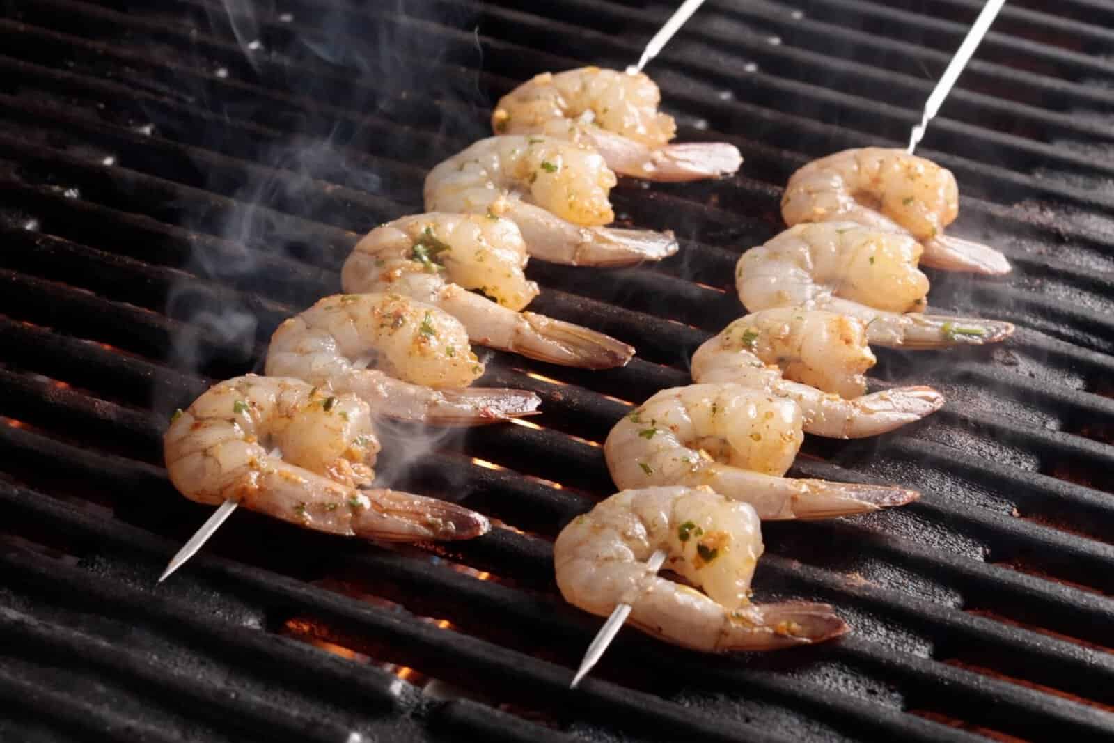 Skewered shrimp cooking on a grill, with visible smoke rising and grill marks starting to form. The shrimp appear seasoned with herbs and spices.