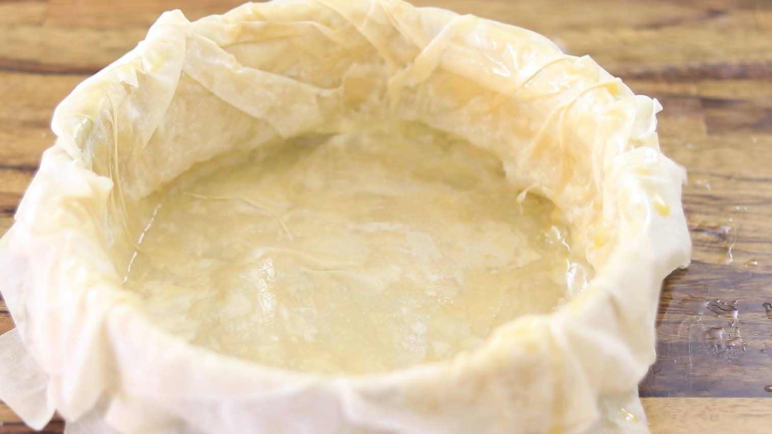 A round baking dish lined with layers of phyllo pastry, brushed with melted butter, sits on a wooden surface, ready to be filled for baking.