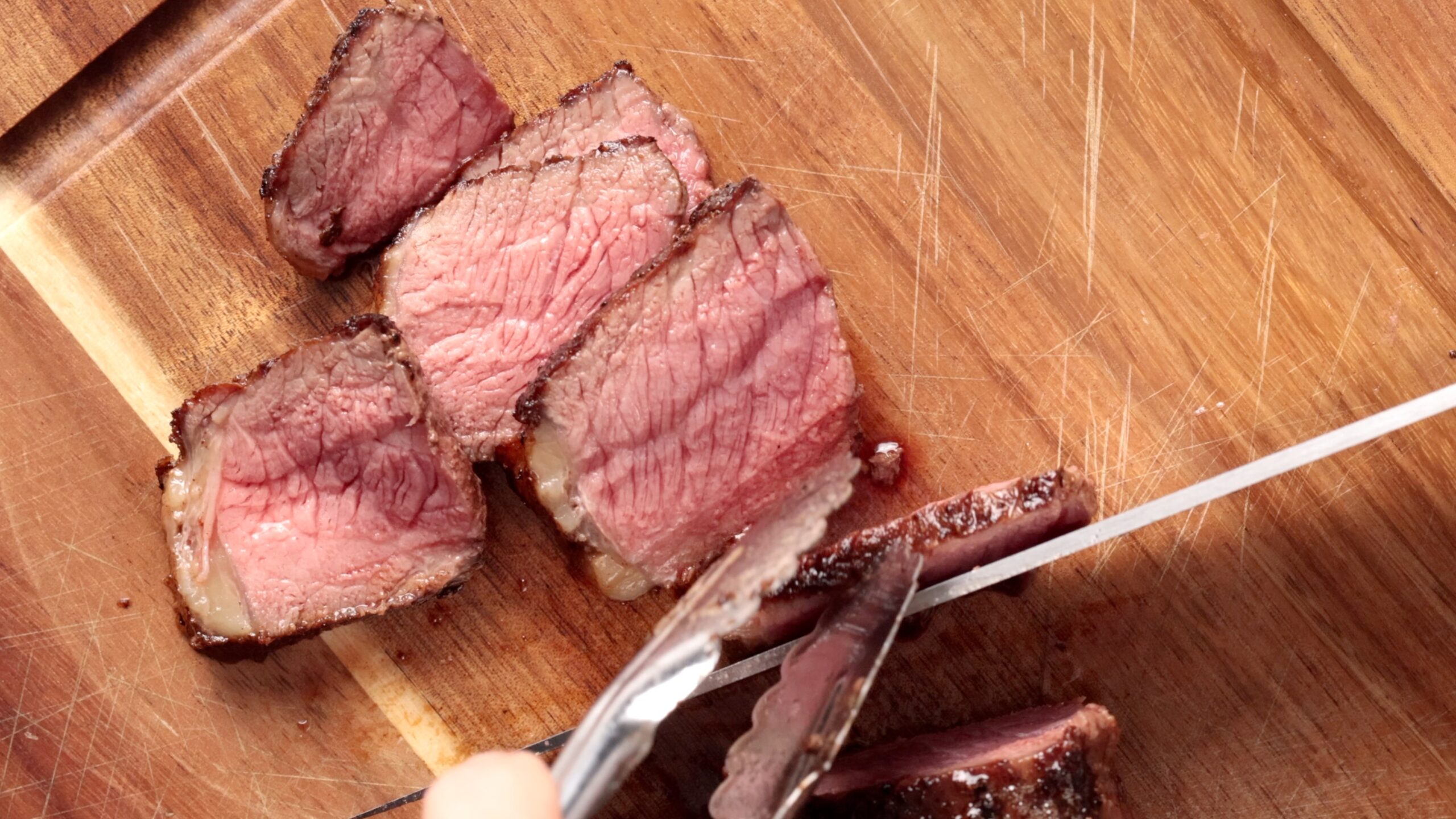 Close-up of a steak being sliced on a wooden cutting board. Several pieces of medium-rare steak are visible, showing a pink, juicy interior. A knife and tongs are being used to cut the meat.