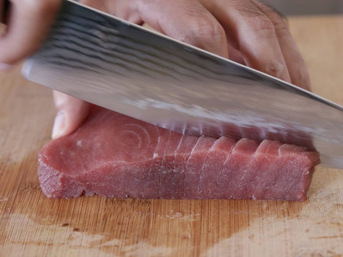 A person slices a piece of raw tuna with a sharp knife on a wooden cutting board.