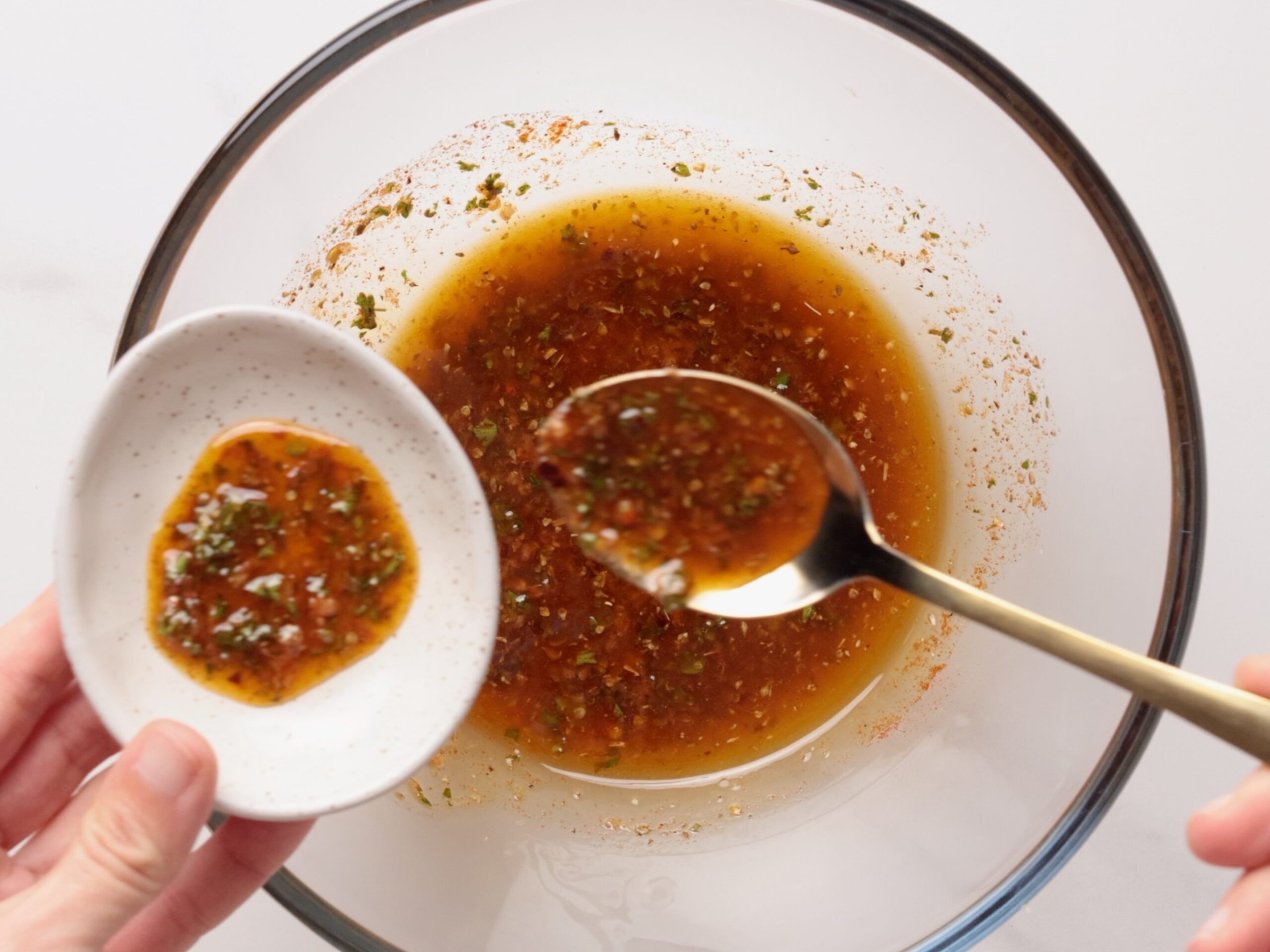 A hand holds a small white dish with a herb and spice marinade for oven baked shrimp skewers, while a spoon scoops more marinade from a glass bowl underneath.