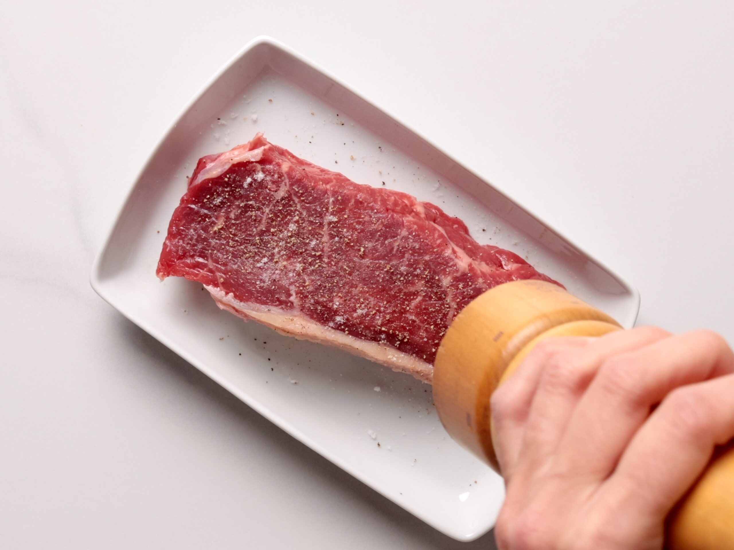 A hand grinds black pepper onto a raw steak that is placed on a small white rectangular plate.
