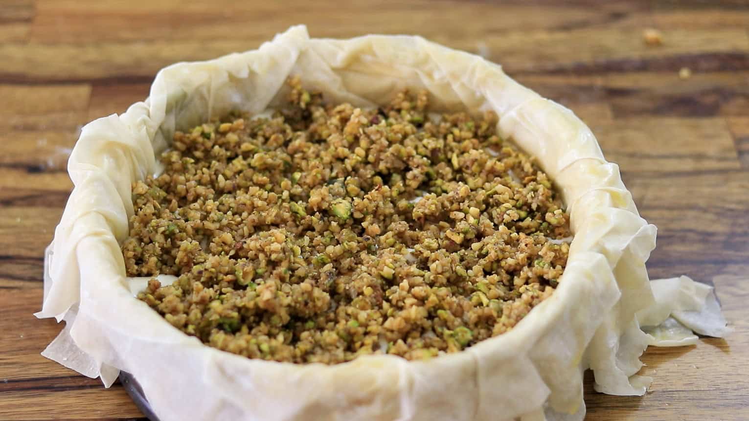 A round baking dish lined with phyllo dough, filled with a chopped nut mixture, sits on a wooden surface, ready to be baked.