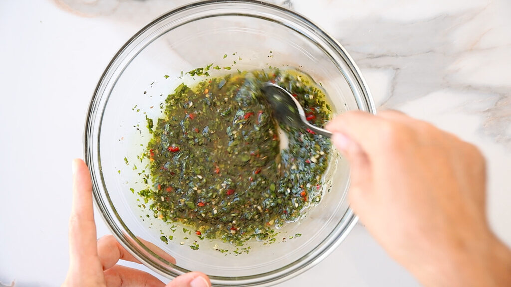 A person stirs a bowl of chimichurri sauce with a spoon, mixing green herbs, red pepper, and oil in a clear glass bowl on a marble countertop.