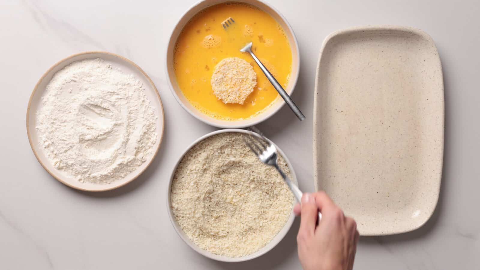 Three bowls: flour, beaten eggs, and breadcrumbs, with a hand using a fork to coat food in breadcrumbs. An empty plate is beside the bowls, all on a light countertop.